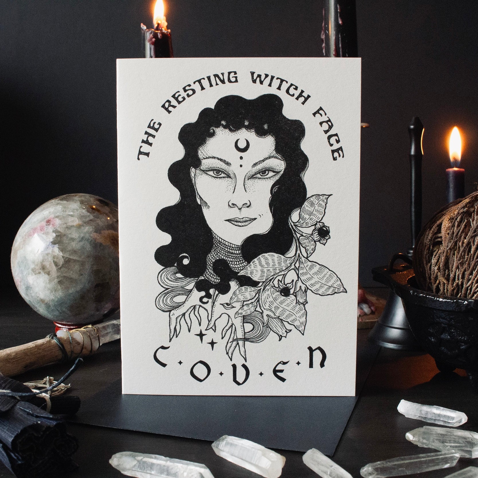 Greeting card with an illustration of a witch's face surrounded by floral elements and the name 'COVEN' below it. The card is displayed on a dark background with candles and crystal stones around.