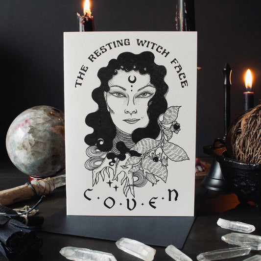Greeting card with an illustration of a witch's face surrounded by floral elements and the name 'COVEN' below it. The card is displayed on a dark background with candles and crystal stones around.